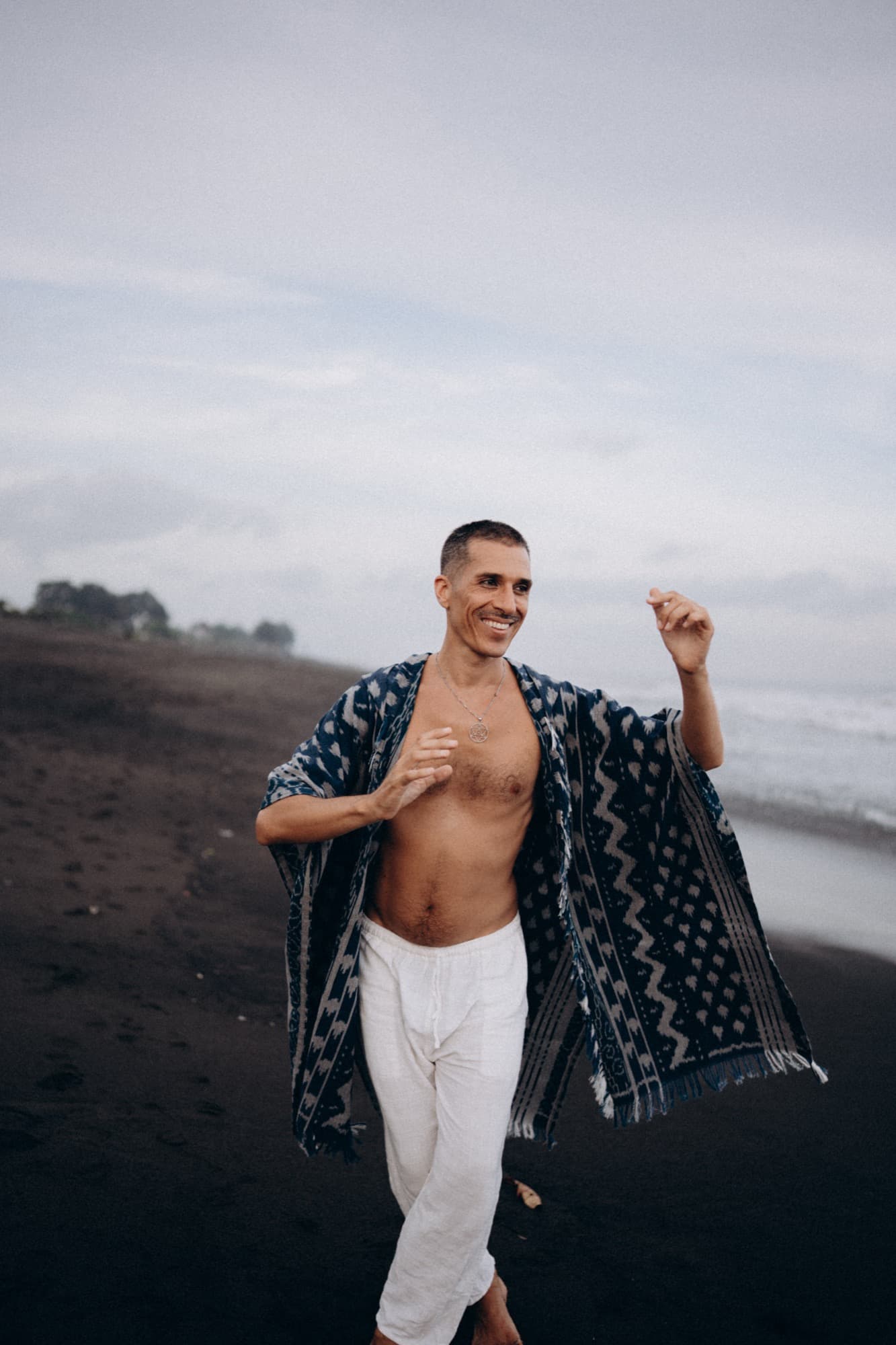 Mysticlio walking and smiling on black sand beside the sea, wearing an indigo ikat robe